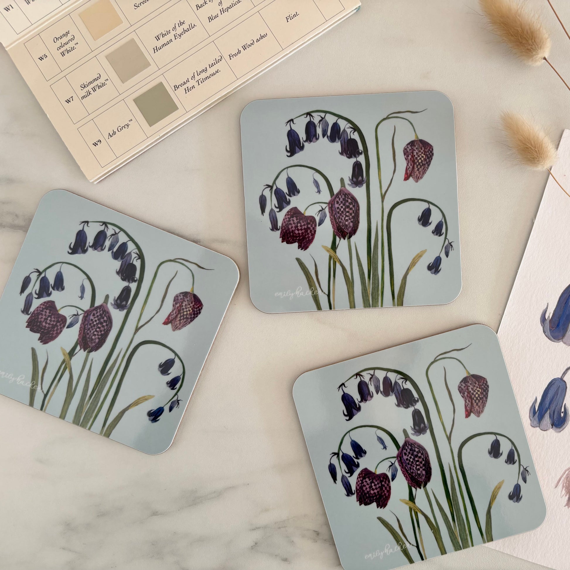 Melamine blue bluebell and snake head fritillary coasters,  next to the hand painted design on a marble background. 