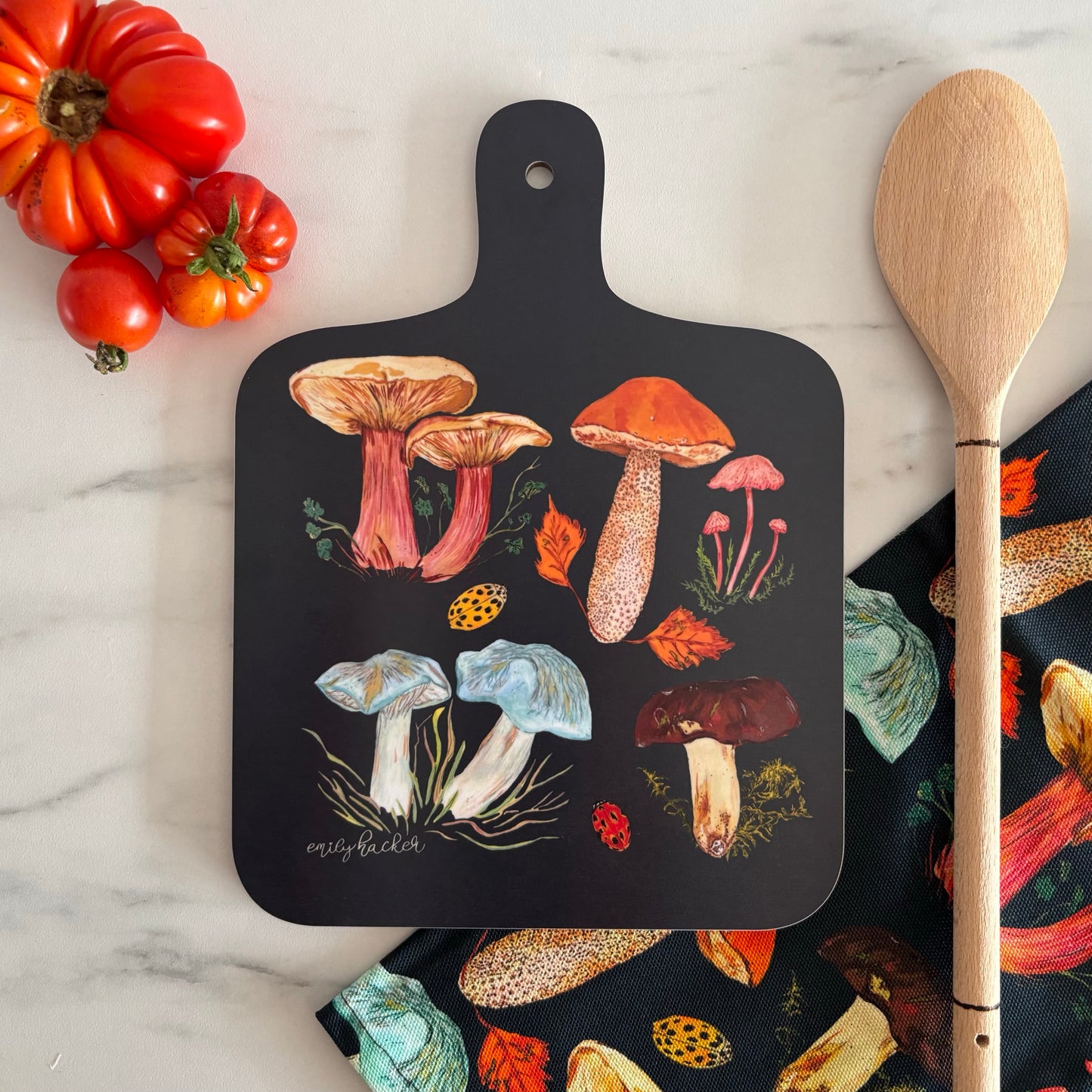 Dark blue cutting board with mushroom design on a marble surface with a wooden spoon and tomatoes.