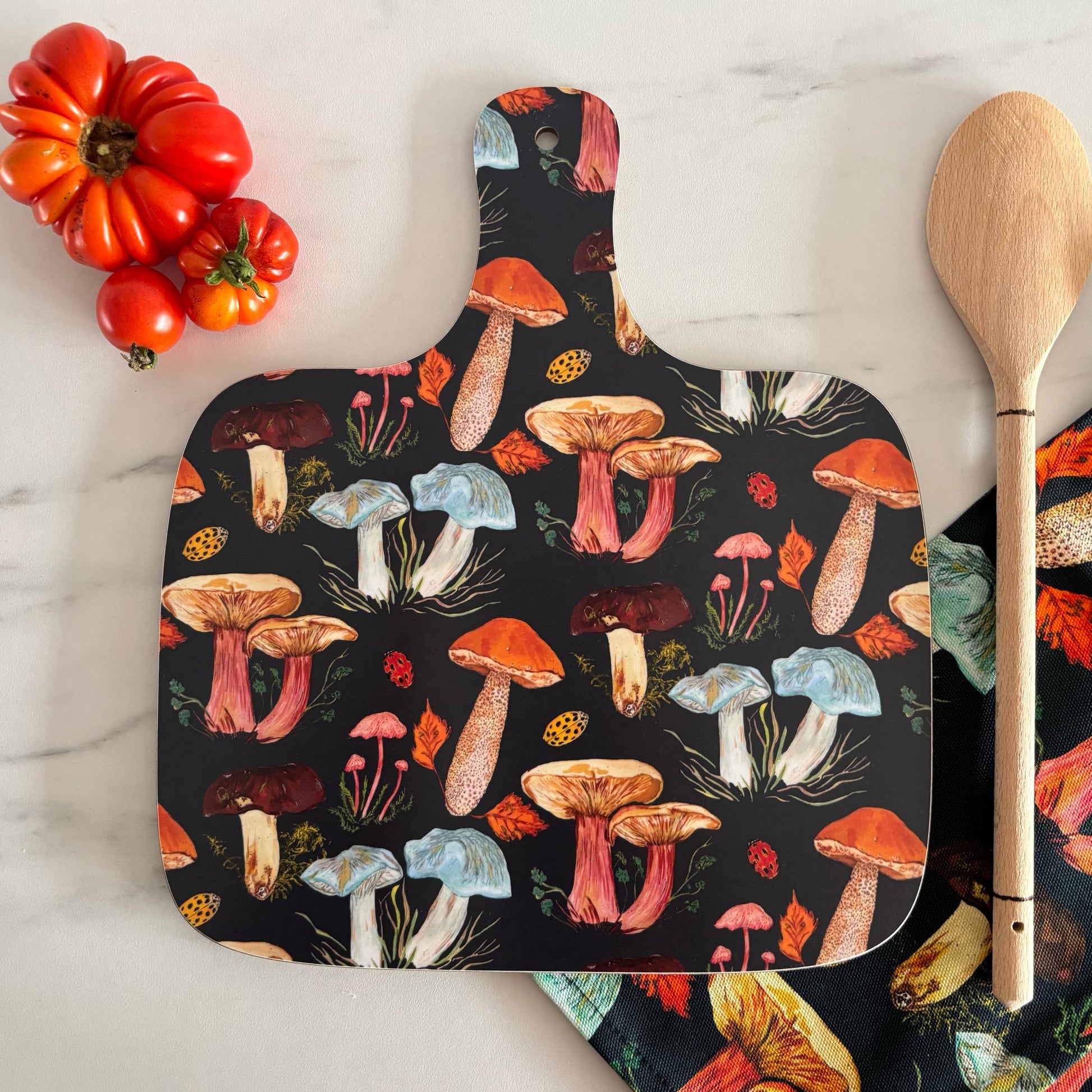 Mushroom chopping board featuring different woodland fungi on a dark ink blue background. The chopping board is photographed next to a autumn mushroom tea towel and tomato.