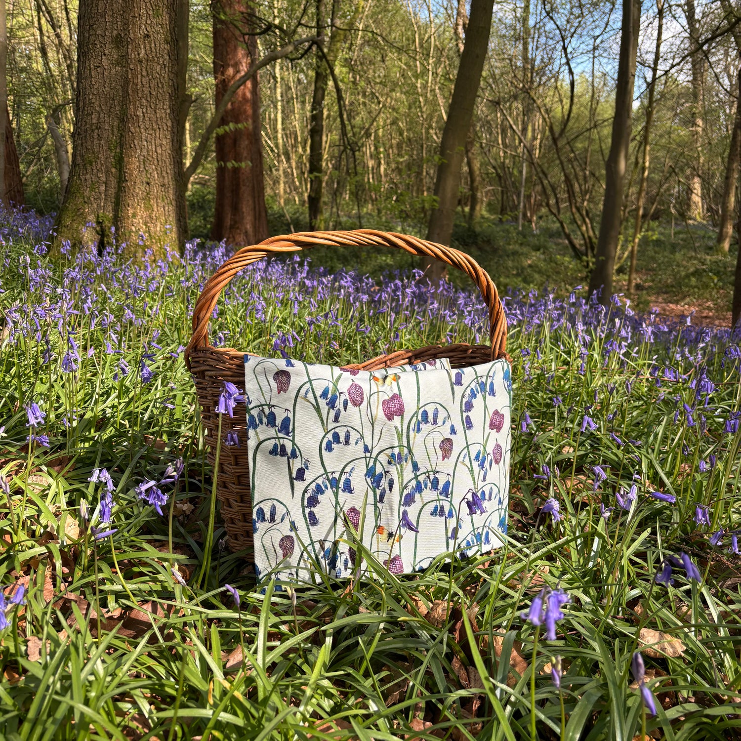 Wicker picnic basket with patterned bluebell tea towel cloth on a grassy area with bluebell flowers and trees.