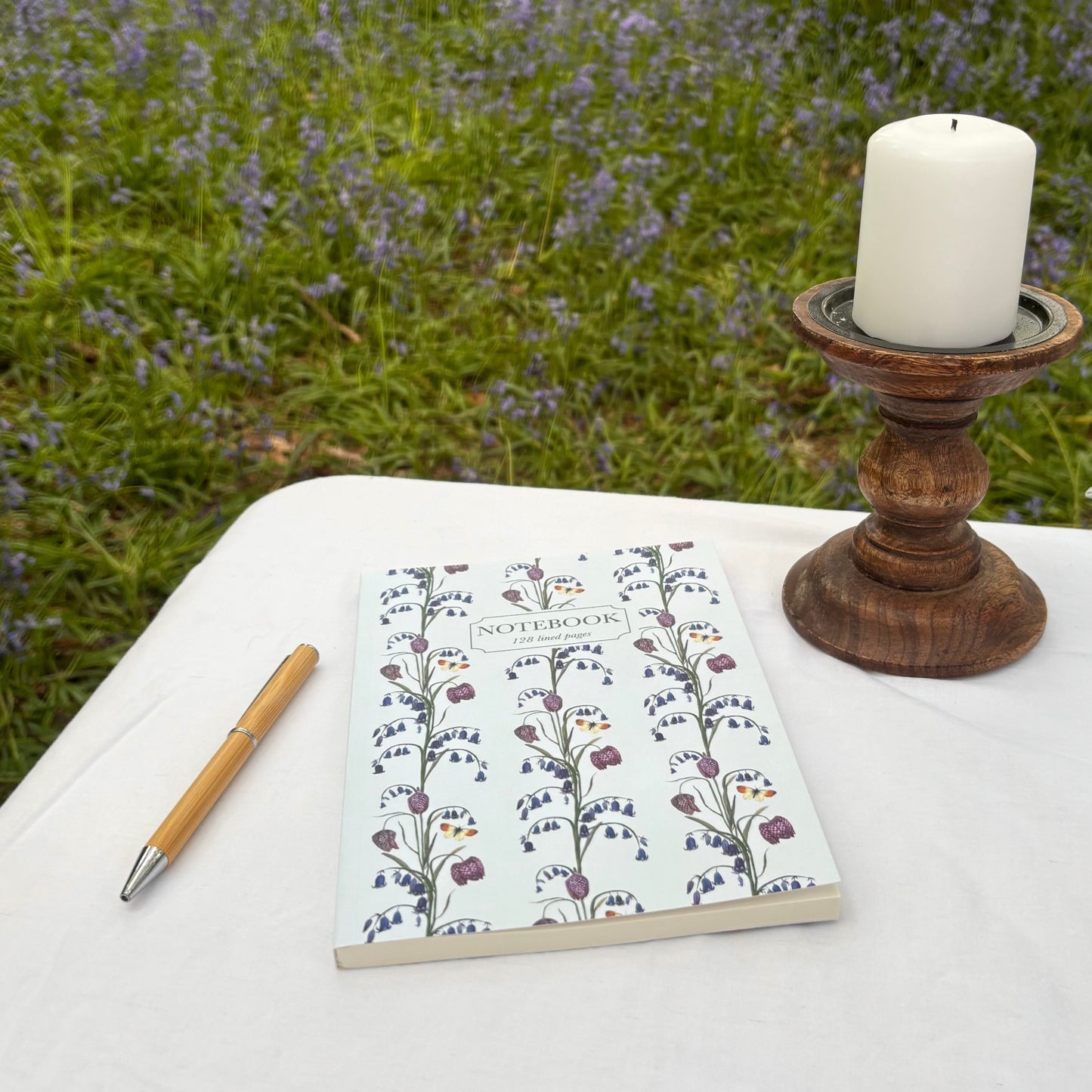 Notebook with British wildflower design, next to eco stationery pen, and candle on a table with a forest floor background