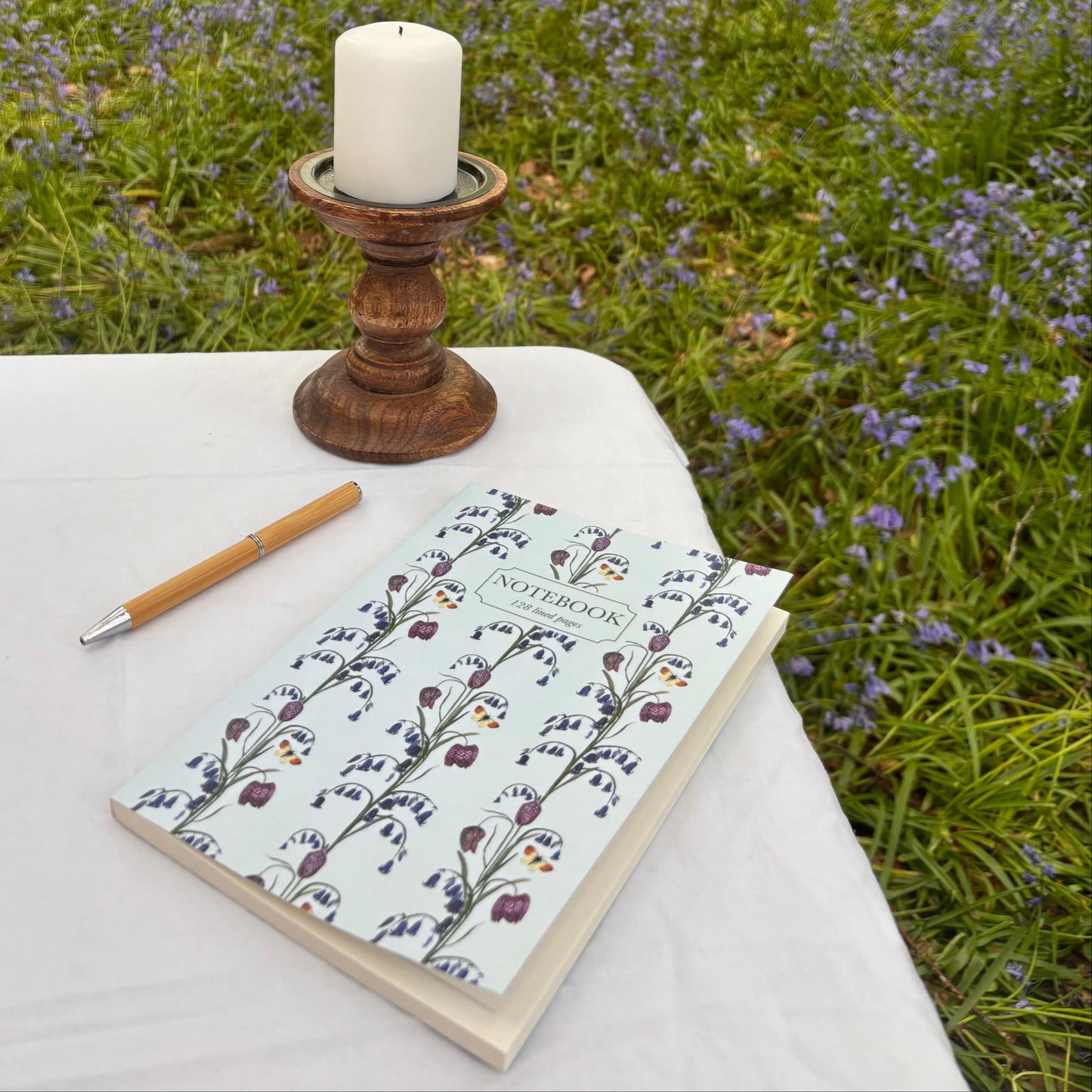 Book with British wildflower bluebell and snakehead design next to a wooden candle holder and pen on a table in a forest with bluebell flowers
