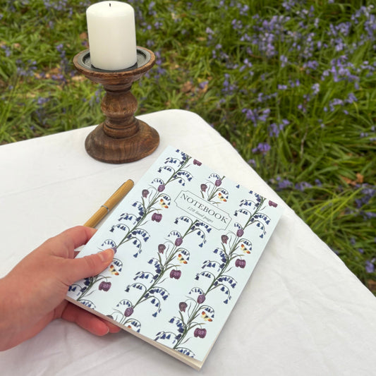 Wildflower Notebook with floral snake head fritillary design held by a hand on a white surface with a candle and pen, against a blurred natural bluebell  background.