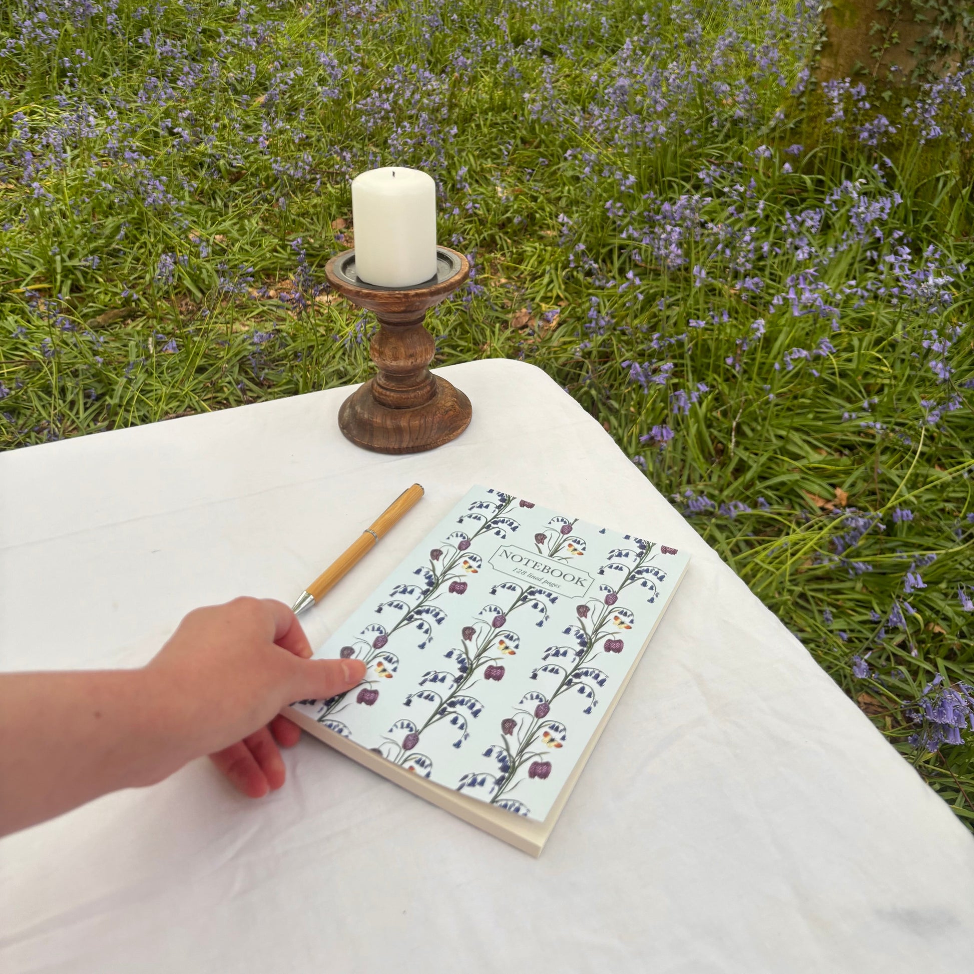 Person holding a wildflower bluebell notebook with a eco stationery pencil on a table in a forest with bluebell flowers and a candle.