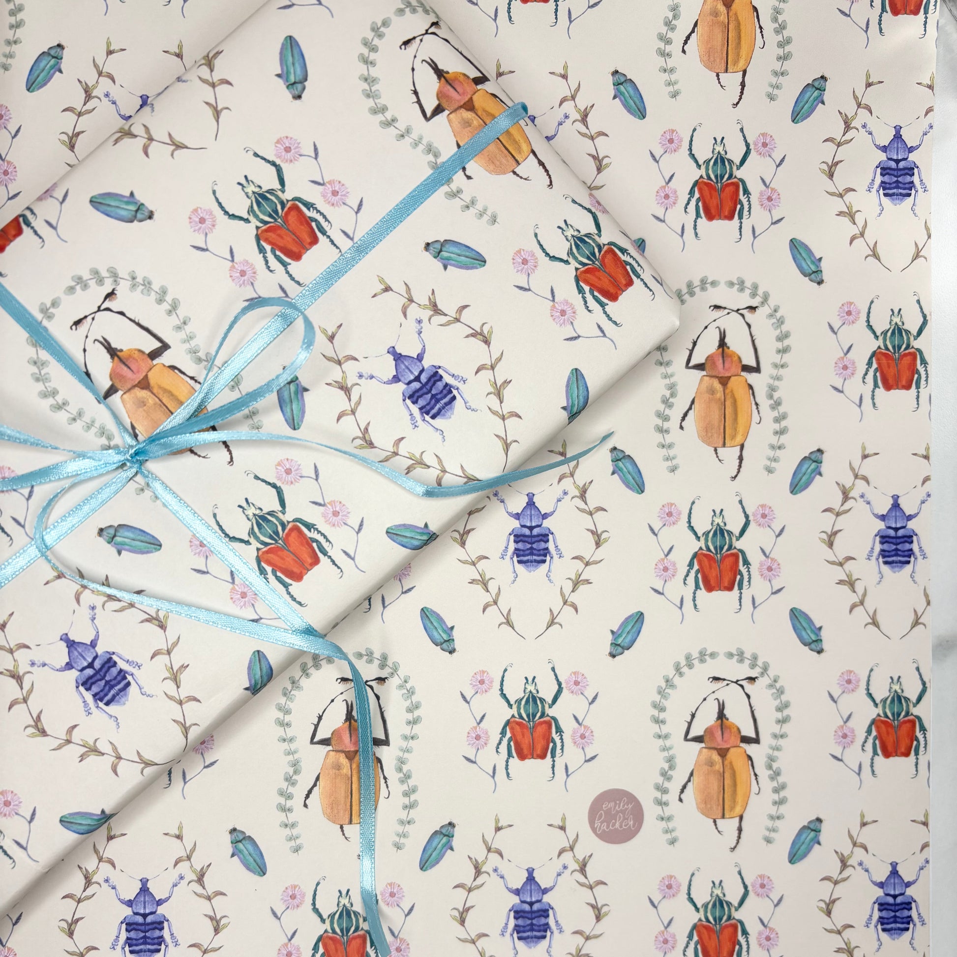 Hand illustrated entomology Gift wrap with beetle and floral pattern with a blue ribbon on a cream background. Illustrations include weevils, Goliath beetles and foliage.