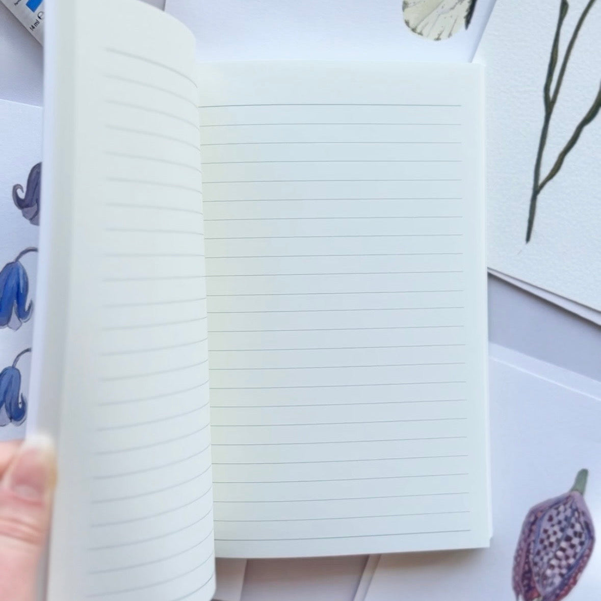 An open lined notebook with lined white pages, held by a hand against a styled flat-lay background of botanical art prints and small stationery items.
