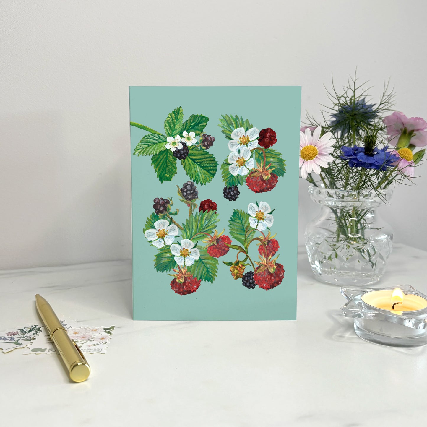 Illustrated strawberry Greeting Card with floral and berry design on a light blue background, placed on a white surface with a pen and small plant in the background.