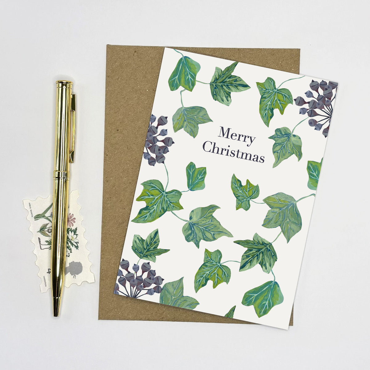 A Christmas greeting card with a green ivy pattern and 'Merry Christmas' printed on it, placed on top of a brown kraft envelope. A gold pen and small decorative paper pieces are arranged beside it on a clean white background.