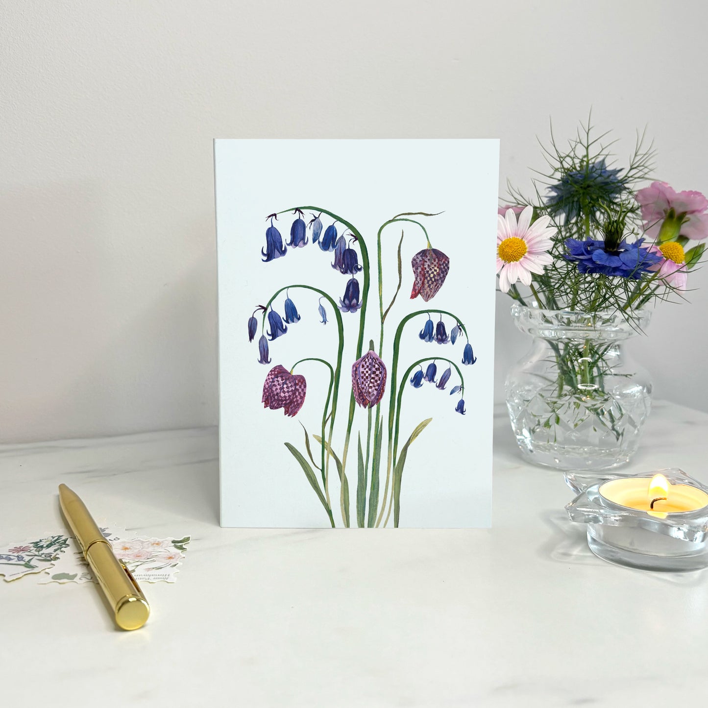 Native wildflower Greeting card with floral bluebell and snake head fritillary design on a white surface with a vase of flowers and candle