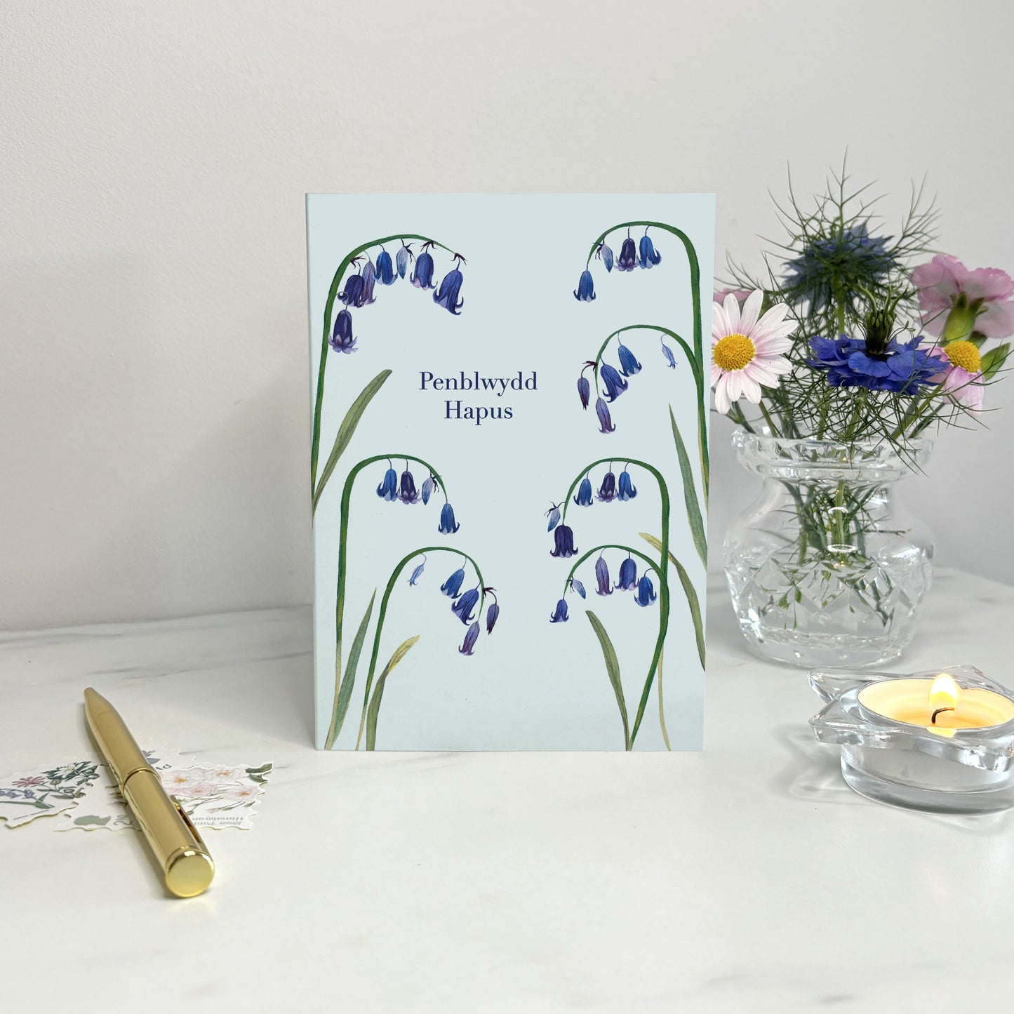 Penblwydd hapus Native wildflower Greeting card with bluebell design on a white surface with a candle and vase.