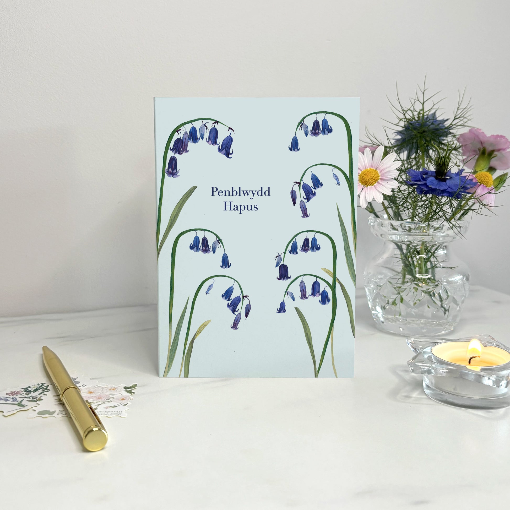 Penblwydd hapus Native wildflower Greeting card with bluebell design on a white surface with a candle and vase.