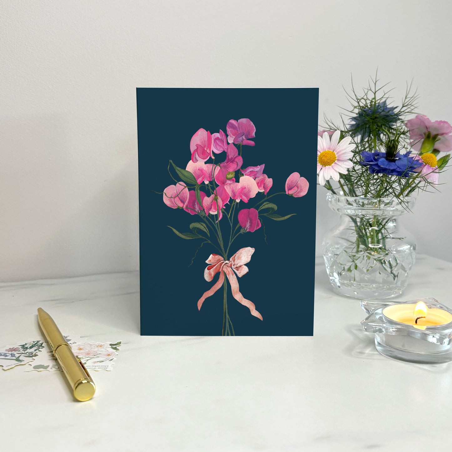Floral wildflower card with pink sweet pea flowers and a ribbon on a blue background, placed on a white surface with a pen and small plant.