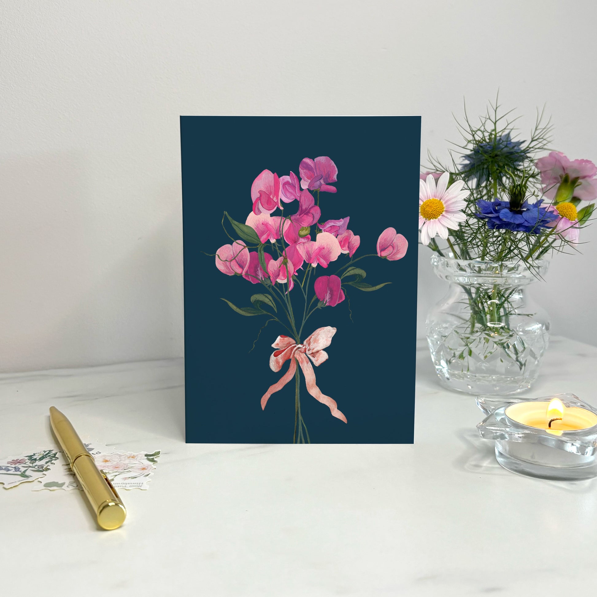 Floral wildflower card with pink sweet pea flowers and a ribbon on a blue background, placed on a white surface with a pen and small plant.