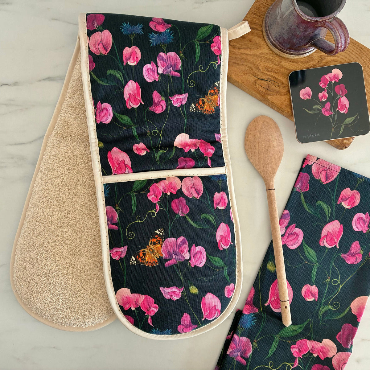 Double oven glove with everlasting sweet pea, blue cornflower and painted lady butterfly design on a dark navy blue background. The oven glove is photographed next to a wooden spoon, matching tea towel, matching coaster on a wooden block.