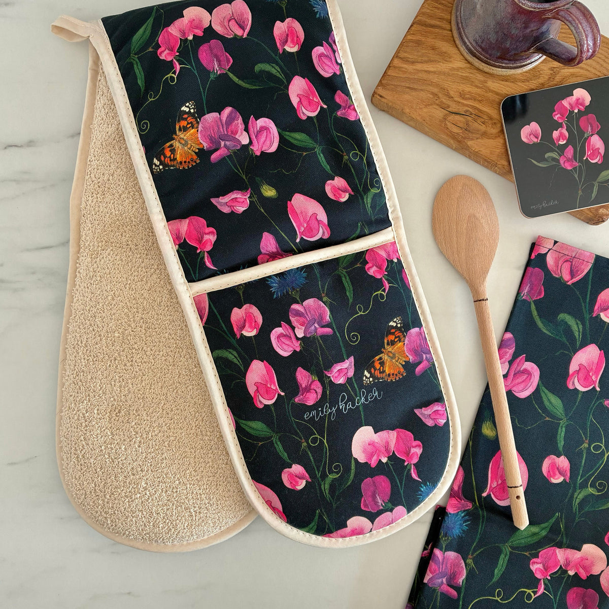 Double oven glove with everlasting sweet pea design on a dark navy blue background.the oven glove is photographed next to a wooden spoon, matching tea towel, matching coaster on a wooden block.