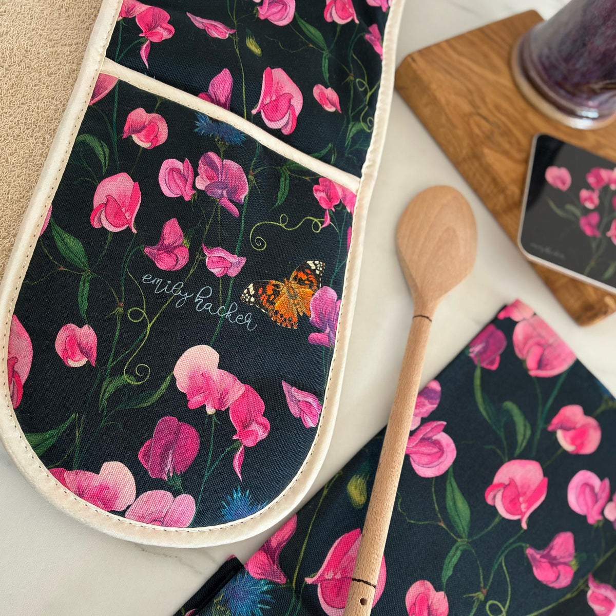 A close-up of a floral kitchen oven glove on a light surface, featuring a hand painted pattern with pink sweet pea flowers and blue cornflowers on a dark background. It’s photographed next to a matching tea towel, coaster and wooden spoon.