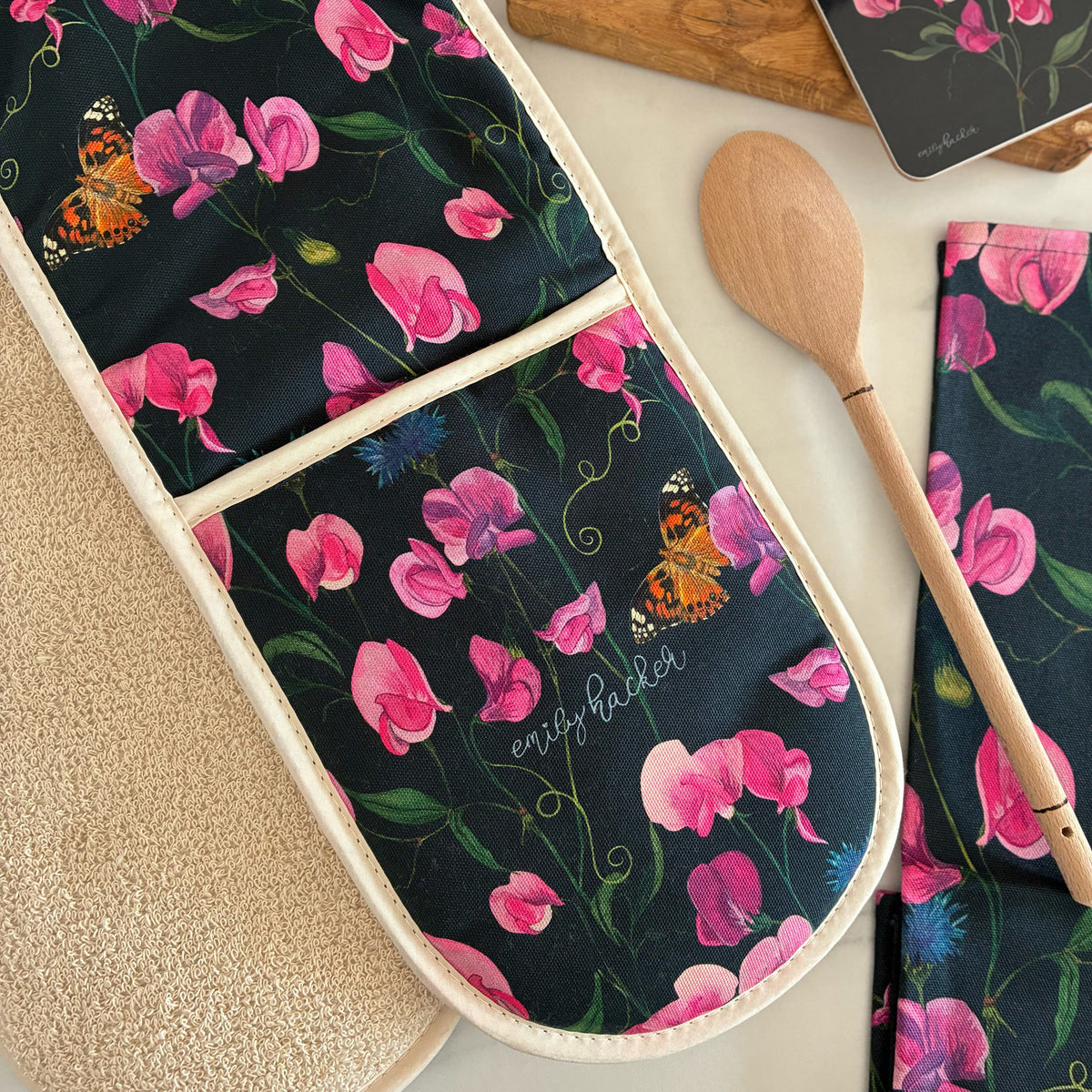 A flat-lay of an oven glove featuring a navy background with pink everlasting sweet pea flowers, blue cornflowers, and painted lady butterflies. The oven glove is photographed on a light marble surface.