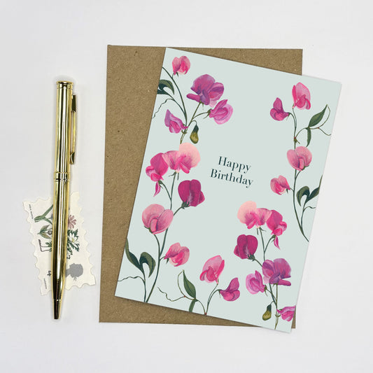 Floral sweet pea birthday card with pink flowers and 'Happy Birthday' text, accompanied by a pen and small postcard on a light gray background.
