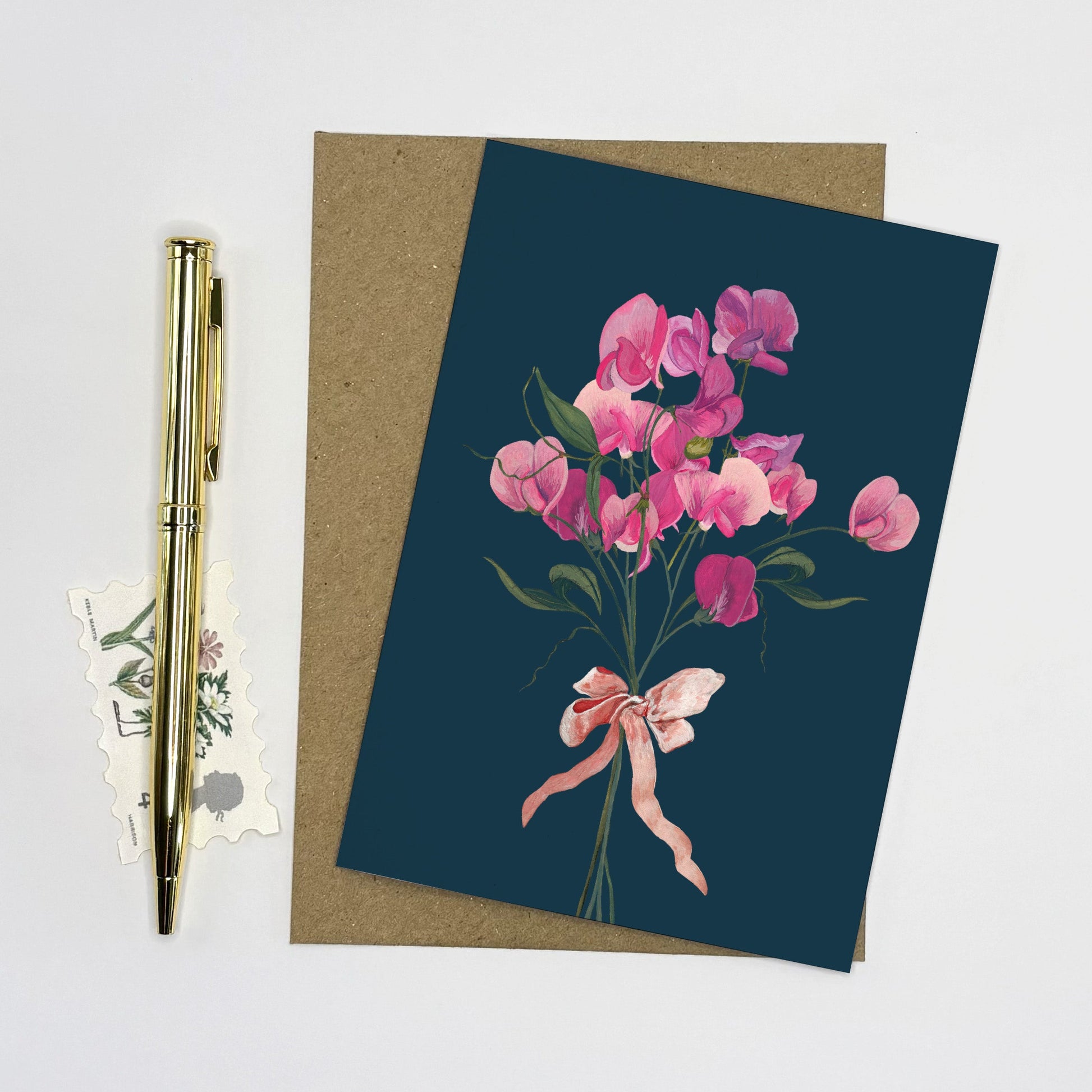 Sweet pea Greeting card with hand painted floral bow design on a dark blue background, placed on a white surface with a pen and stickers.