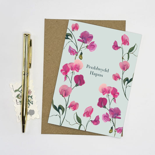 Sweet pea Floral birthday card with pink flowers and 'Happy Birthday' text, accompanied by a pen and small postcard on a light gray background.