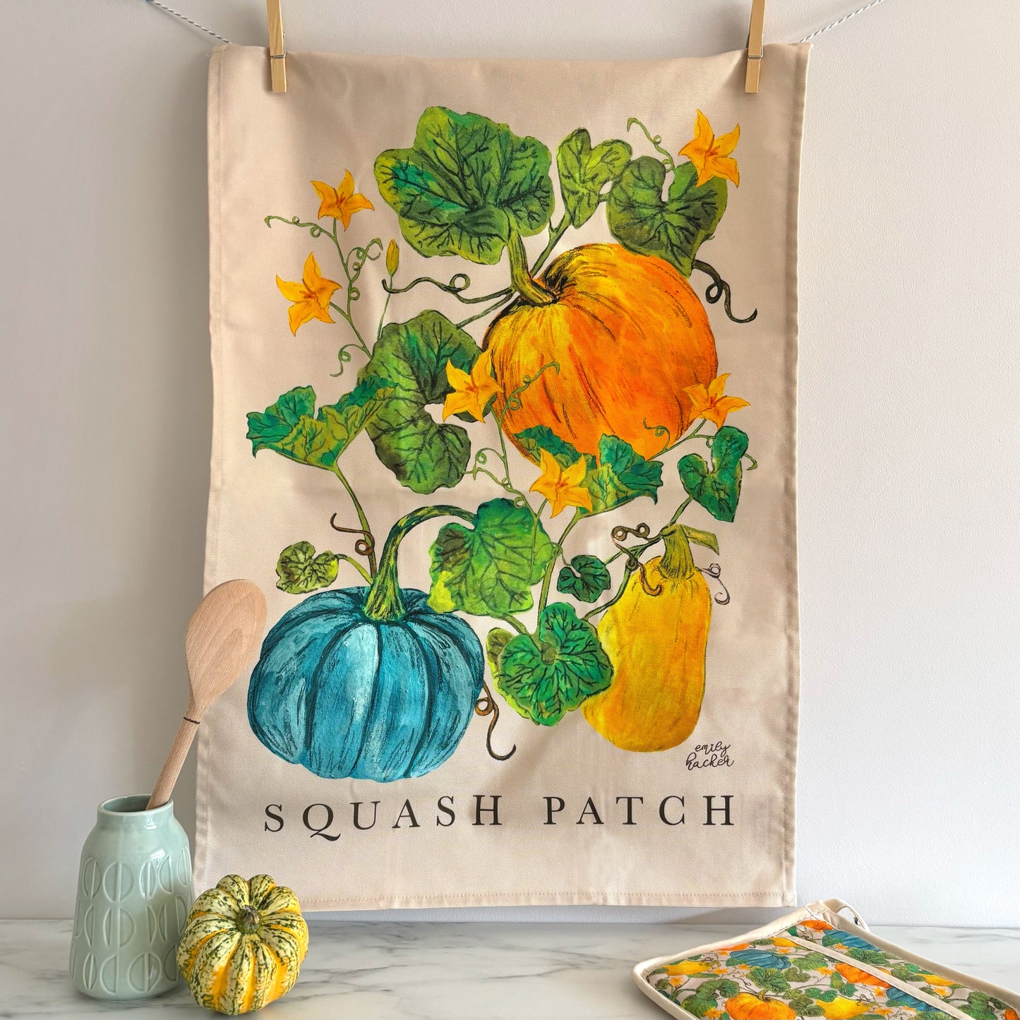 Tea towel with squash patch design, with small and large orange, heritage blue pumpkins and yellow squash. The tea towel is hung above a marble surface where there is a wooden spoon pumpkin pot grab and a gourd.