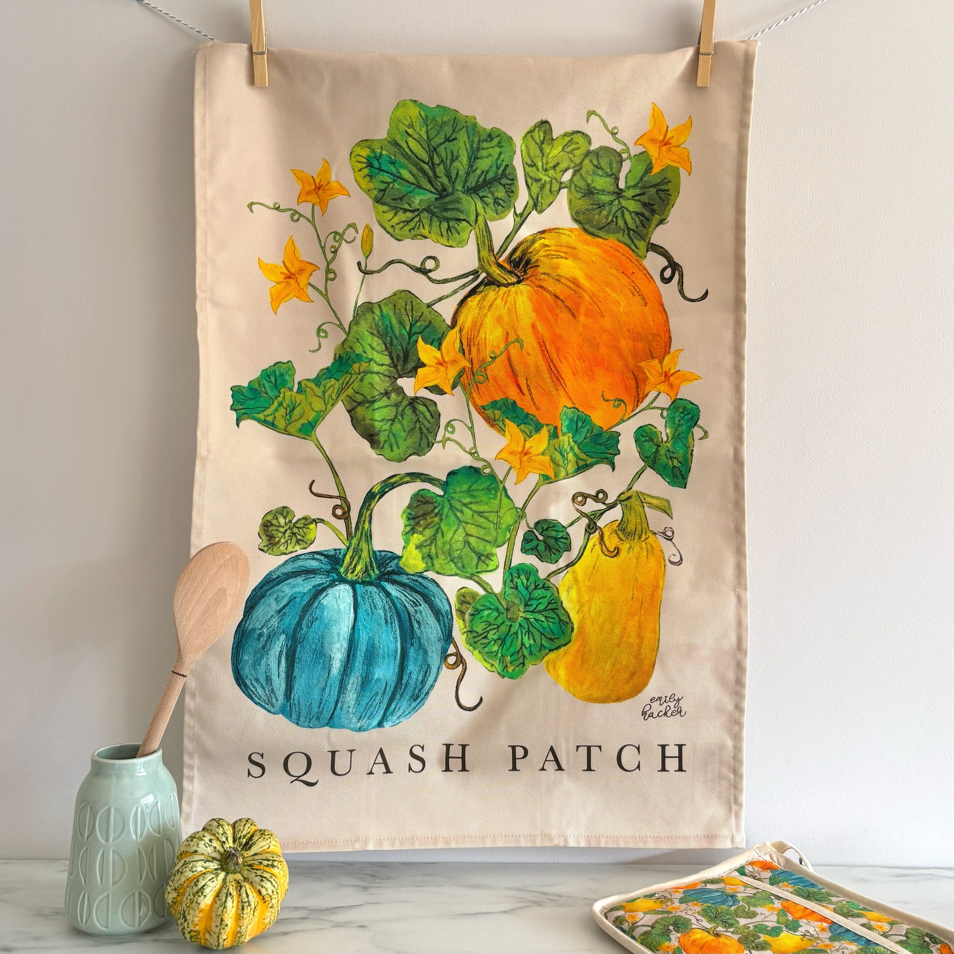 Tea towel with squash patch design, with small and large orange, heritage blue pumpkins and yellow squash. The tea towel is hung above a marble surface where there is a wooden spoon pumpkin pot grab and a gourd.