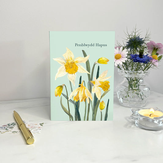 Penblwydd hapus  Welsh Birthday card with daffodils on a white surface with a vase of flowers and pen.