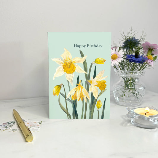 Daffodil Birthday card with daffodils on a white surface with a vase of flowers and pen.