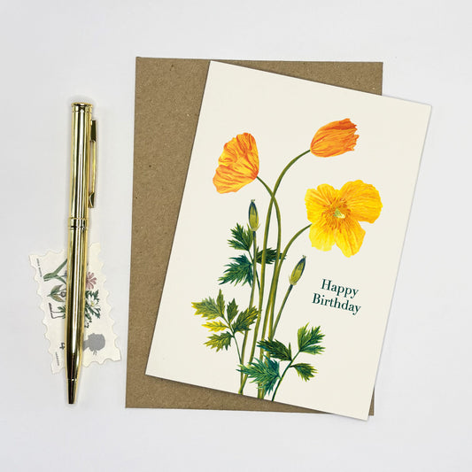 Greeting card with yellow Welsh poppy wildflowers and 'Happy Birthday' text, accompanied by a pen and small postcard on a light gray background.
