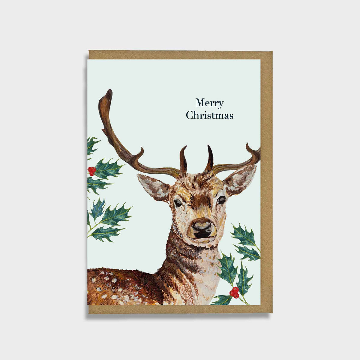 A rectangular Christmas greeting card with a brown deer illustration, green holly leaves, and red berries on a pale background, with the text 'Merry Christmas' near the top. The card is shown standing upright against a light gray background.
